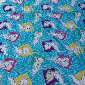 Op de afbeelding: Een turquoise stof met een herhaald patroon van Casper het Vriendelijke Spook. Elke Casper is omlijst in een kleurrijke ruitvorm, tegen een wervelende blauwe achtergrond. De ruiten zijn geel, paars, groen en roze.