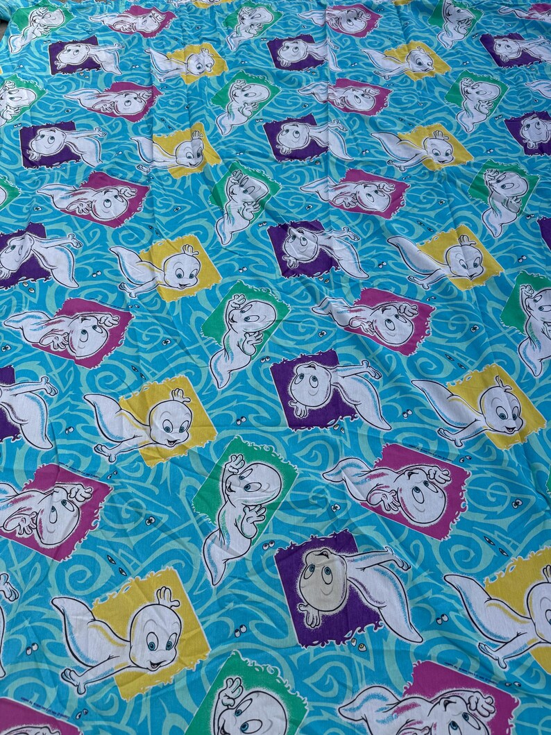 Op de afbeelding: Een turquoise stof met een herhaald patroon van Casper het Vriendelijke Spook. Elke Casper is omlijst in een kleurrijke ruitvorm, tegen een wervelende blauwe achtergrond. De ruiten zijn geel, paars, groen en roze.