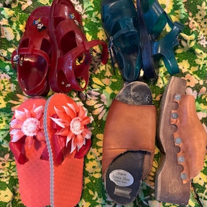 May include: A collection of vintage footwear, including red jelly sandals with cartoon characters, teal plastic sandals, orange flip-flops with flower embellishments, and brown leather clogs. The shoes are arranged on a floral patterned fabric.