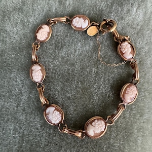 May include: A gold-toned link bracelet featuring six oval-shaped cameos, each displaying a white profile relief against a light brown background. The bracelet includes a circular spring ring clasp and a small round metal tag attached near the closure.
