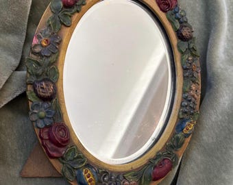 May include: Oval-shaped decorative mirror with a detailed frame. The frame is adorned with colorful, three-dimensional fruit, flowers, and leaves. The mirror has a light brown wooden stand.