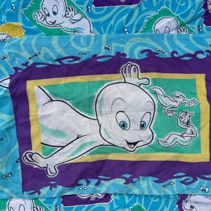 Op de afbeelding: Een Casper the Friendly Ghost kussensloop. De kussensloop heeft een grote afbeelding van Casper in een paarse, gele en groene lijst. De achtergrond is lichtblauw met Casper spookfiguren en golvende lijnen.
