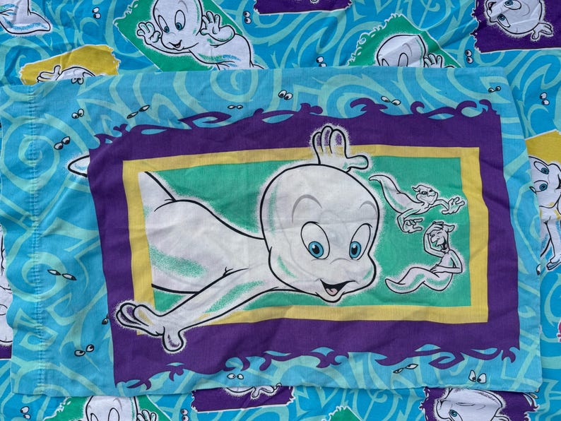 Op de afbeelding: Een Casper the Friendly Ghost kussensloop. De kussensloop heeft een grote afbeelding van Casper in een paarse, gele en groene lijst. De achtergrond is lichtblauw met Casper spookfiguren en golvende lijnen.