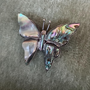 May include: A butterfly-shaped brooch featuring iridescent abalone shell inlays on the right wings and smooth, pearlescent shell inlays on the left wings. The body is crafted from a metallic frame with two thin antennae extending from the head.