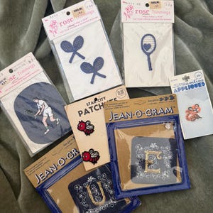 May include: A collection of vintage iron-on patches and appliques, including tennis rackets, a tennis player, bees, and initial letters. The patches are in various colors, including blue, red, and gold, and are packaged in clear plastic.