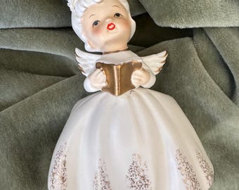 May include: A ceramic figurine of an angel with white hair, a floral headpiece, and small wings. The angel wears a white dress with gold accents and holds a gold book. The facial features include painted red lips and dark eyes.