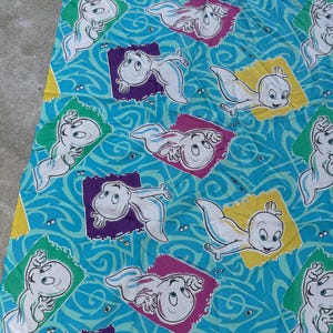 Op de afbeelding: Een turquoise stof met een herhaald patroon van het stripfiguur Casper het Vriendelijke Spook. Elke Casper staat in een kleurrijk vierkant, tegen een wervelende blauwe achtergrond.