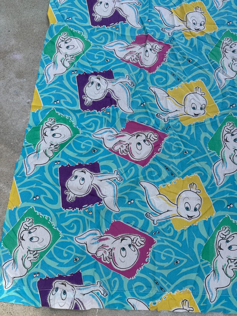 Op de afbeelding: Een turquoise stof met een herhaald patroon van het stripfiguur Casper het Vriendelijke Spook. Elke Casper staat in een kleurrijk vierkant, tegen een wervelende blauwe achtergrond.