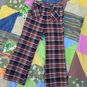 May include: A pair of vintage plaid pants in a classic tartan pattern. The pants feature a mix of red, navy, yellow, and brown hues. The pants have a high waist and a flared leg. The pants are displayed on a colorful patchwork quilt.