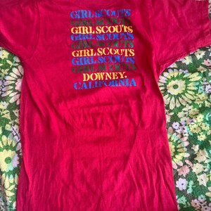 May include: A red t-shirt with the words "GIRL SCOUTS" stacked vertically in multiple colors. Below the text is "DOWNEY, CALIFORNIA" in blue. The shirt has short sleeves and a crew neck.