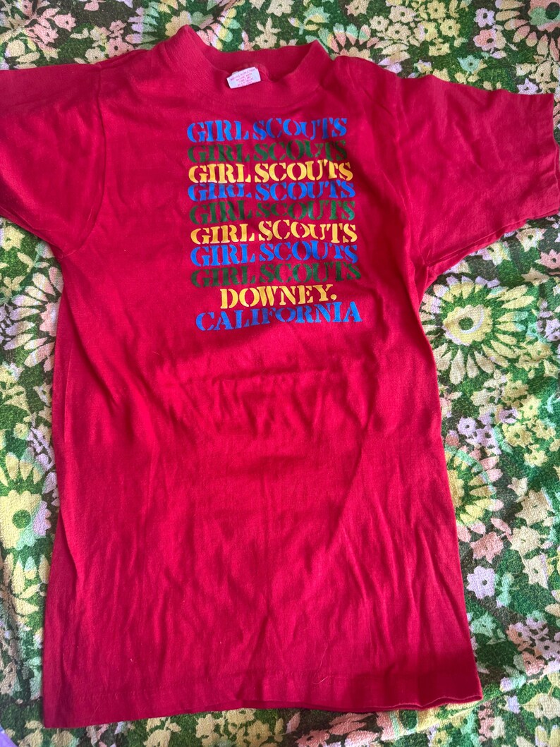 May include: A red t-shirt with the words "GIRL SCOUTS" stacked vertically in multiple colors. Below the text is "DOWNEY, CALIFORNIA" in blue. The shirt has short sleeves and a crew neck.