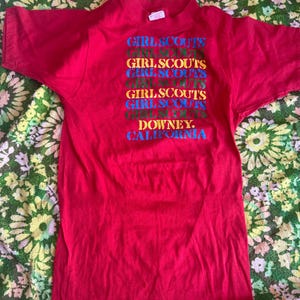 May include: A red t-shirt with the words "GIRL SCOUTS" stacked vertically in multiple colors. Below the text is the word "DOWNEY, CALIFORNIA". The shirt is made of a soft material.