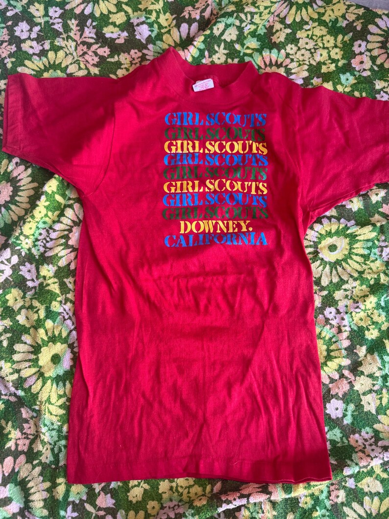 May include: A red t-shirt with the words "GIRL SCOUTS" stacked vertically in multiple colors. Below the text is the word "DOWNEY, CALIFORNIA". The shirt is made of a soft material.