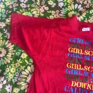 May include: A red t-shirt with the text "GIRLSCO" stacked vertically in multiple colors. The text "DOWNTOWN CALIFORNIA" is below the stacked text. The shirt is made of a soft material.
