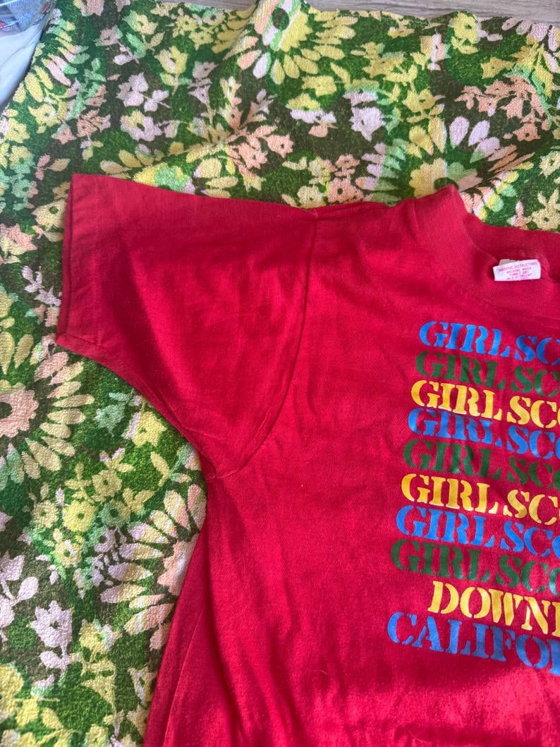 May include: A red t-shirt with the text "GIRLSCO" stacked vertically in multiple colors. The text "DOWNTOWN CALIFORNIA" is below the stacked text. The shirt is made of a soft material.