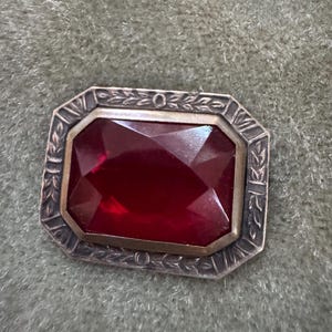 May include: A rectangular brooch with a large, faceted, deep red gemstone. The gem is set within a silver-toned frame with a gold-toned inner border. The frame features a decorative leaf and circle design.