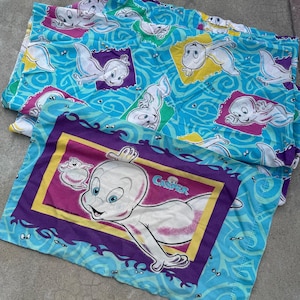 Op de afbeelding: Twee Casper the Friendly Ghost kussenslopen. E&eacute;n kussensloop heeft een grote Casper-afbeelding met het woord "Casper" in een paarse en gele lijst. De andere kussensloop heeft een blauwe achtergrond met Casper-afbeeldingen in kleurrijke vierkanten.