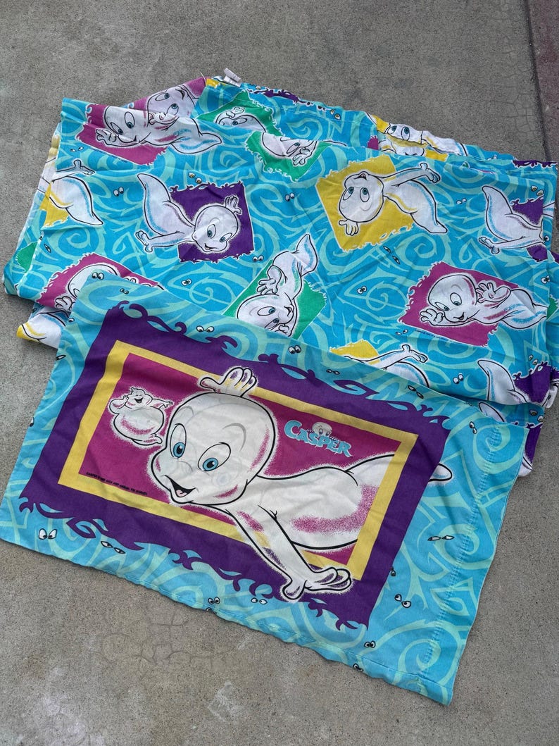 Op de afbeelding: Twee Casper the Friendly Ghost kussenslopen. E&eacute;n kussensloop heeft een grote Casper-afbeelding met het woord "Casper" in een paarse en gele lijst. De andere kussensloop heeft een blauwe achtergrond met Casper-afbeeldingen in kleurrijke vierkanten.