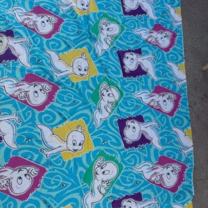 Op de afbeelding: Een lichtblauwe stof met een herhaald patroon van Casper, het vriendelijke spookje. Casper wordt in verschillende poses getoond binnen kleurrijke, rechthoekige kaders tegen een wervelende achtergrond.