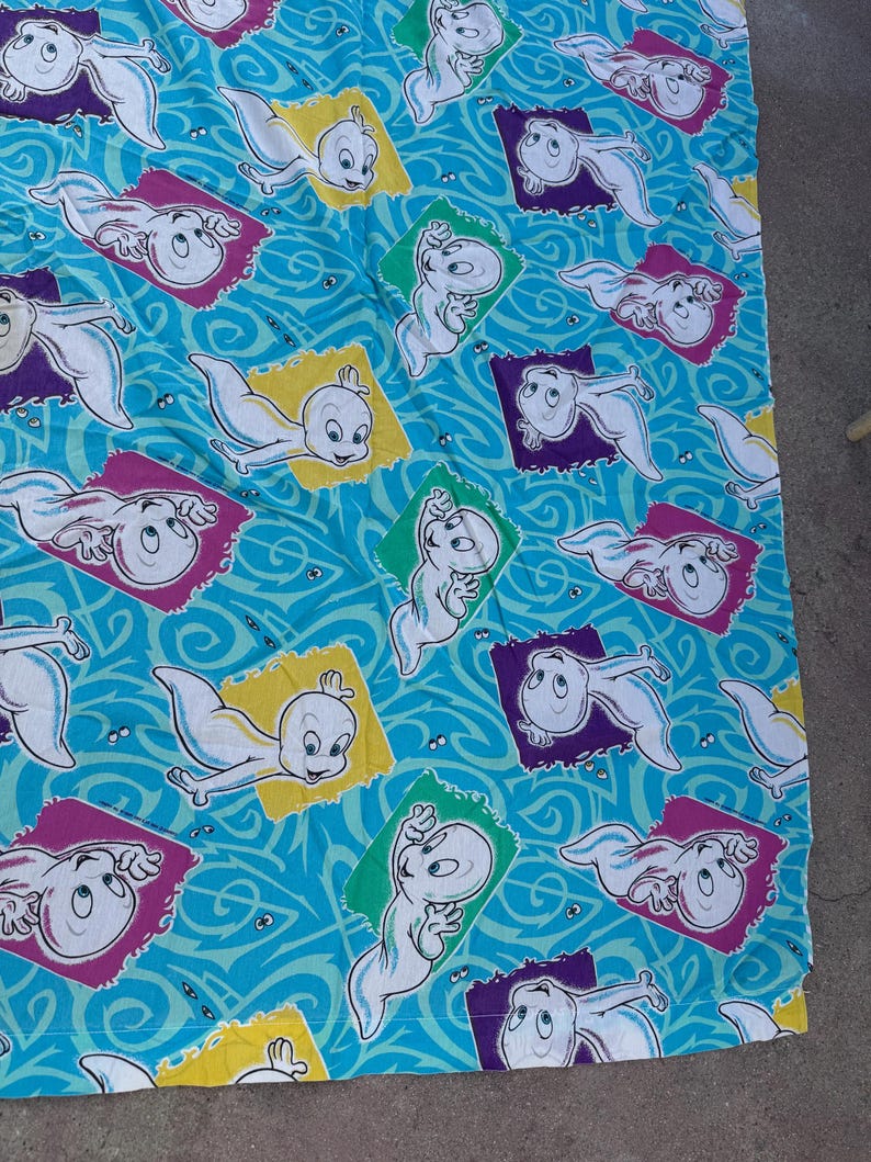 Op de afbeelding: Een lichtblauwe stof met een herhaald patroon van Casper, het vriendelijke spookje. Casper wordt in verschillende poses getoond binnen kleurrijke, rechthoekige kaders tegen een wervelende achtergrond.