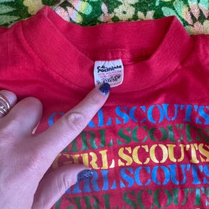 May include: Red t-shirt with the words "GIRL SCOUTS" in a repeating pattern of blue, green, yellow, and red. The shirt has a crew neck and a tag that reads "50% Polyester 50% Cotton".