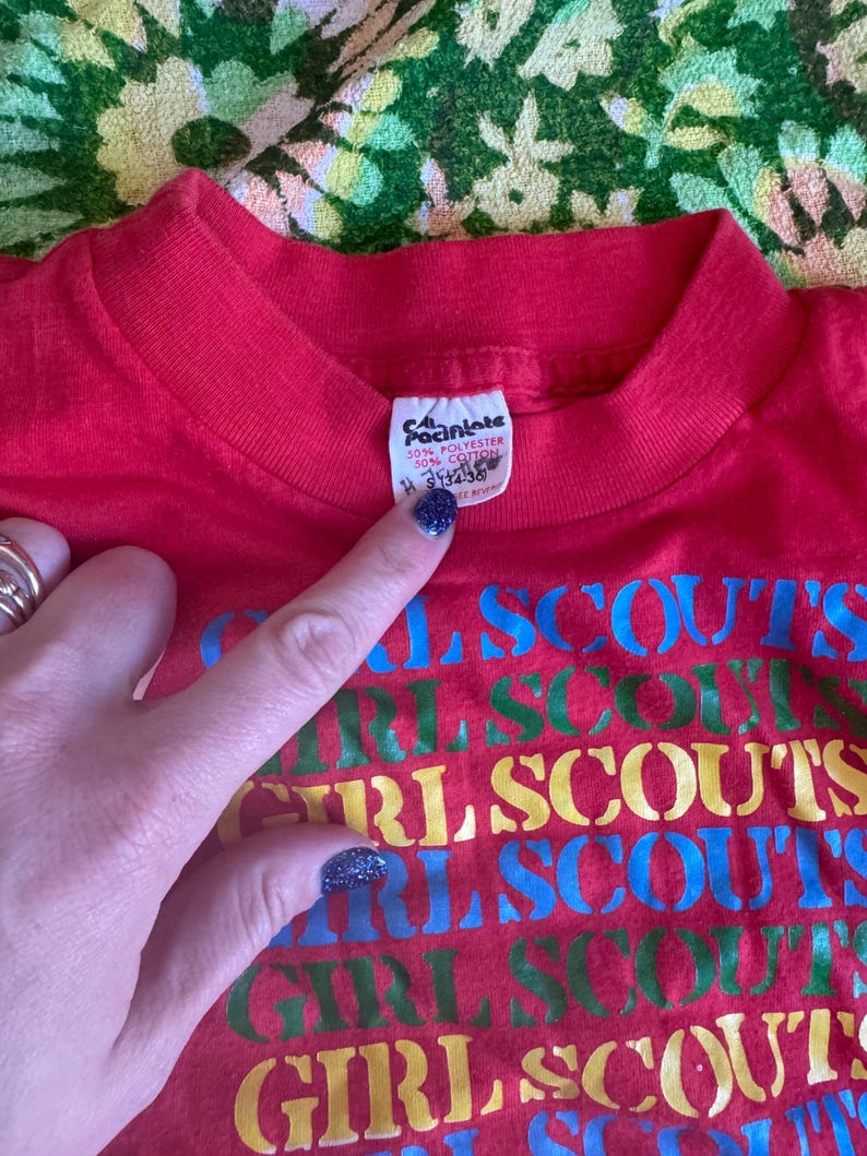 May include: Red t-shirt with the words "GIRL SCOUTS" in a repeating pattern of blue, green, yellow, and red. The shirt has a crew neck and a tag that reads "50% Polyester 50% Cotton".