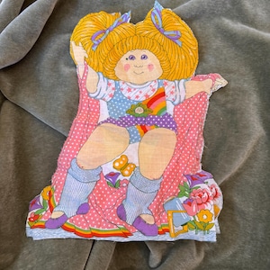 May include: A vintage Cabbage Patch Kids paper doll cutout. The doll has yellow hair with blue bows, wearing a polka dot outfit with a rainbow and butterfly accents. The doll is sitting on a floral design.