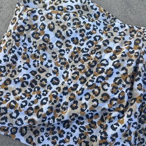 May include: A fabric textile featuring a repeating leopard print pattern in shades of black, dark brown, and tan against a white background. The material is folded, showing the texture and the consistent distribution of the animal print design.