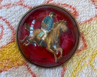Könnte beinhalten: Runde Brosche im antiken Stil mit rotem Hintergrund und detailliertem Bild einer Person, die ein Pferd reitet. Der Reiter trägt eine blaue Jacke und das Pferd ist braun. Die Brosche hat einen goldfarbenen Metallrahmen.