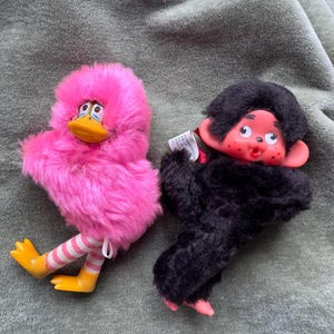 May include: Two vintage plush toys: a pink duck with yellow feet and striped legs, and a black monkey with a red face. The duck has a fluffy pink body and a yellow beak. The monkey has a black fur body and a red face.