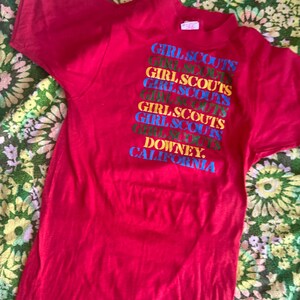 May include: A red t-shirt featuring the text "GIRL SCOUTS" stacked vertically in multiple colors. The text "DOWNEY, CALIFORNIA" is at the bottom. The shirt is made of a soft material.