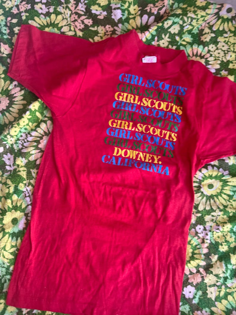 May include: A red t-shirt featuring the text "GIRL SCOUTS" stacked vertically in multiple colors. The text "DOWNEY, CALIFORNIA" is at the bottom. The shirt is made of a soft material.