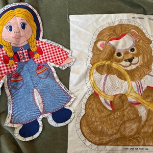 May include: Two fabric cutouts for sewing projects. One is a girl with blonde braids, wearing a red and white checkered top and blue overalls. The other is a lion holding a tennis racket. Both have instructions.