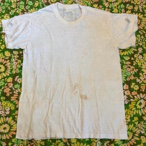 May include: A vintage white t-shirt with a crew neck and short sleeves. The shirt shows signs of wear, including stains and discoloration. The background is a floral patterned fabric in shades of green, yellow, and brown.