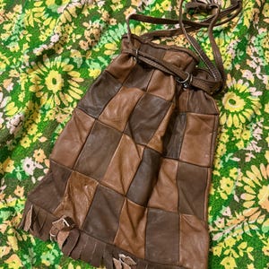 May include: A brown leather patchwork shoulder bag with a drawstring closure and a long strap. The bag features a checkerboard pattern of various shades of brown leather and a fringed bottom. The bag is displayed on a floral patterned fabric.