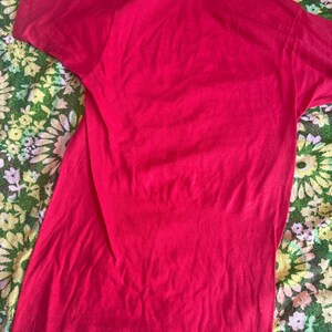 May include: A solid red t-shirt with short sleeves. The shirt is made of a soft material and is slightly wrinkled. The t-shirt is displayed on a floral patterned surface.