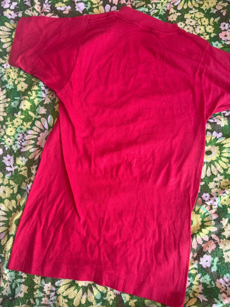 May include: A solid red t-shirt with short sleeves. The shirt is made of a soft material and is slightly wrinkled. The t-shirt is displayed on a floral patterned surface.