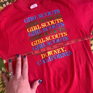 May include: A red t-shirt with the words "GIRL SCOUTS" in a stacked, colorful design. The text "DOWNEY, CALIFORNIA" is printed below. The shirt is being measured with a tape measure.