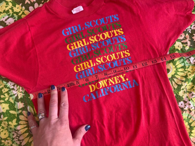 May include: A red t-shirt with the words "GIRL SCOUTS" in a stacked, colorful design. The text "DOWNEY, CALIFORNIA" is printed below. The shirt is being measured with a tape measure.