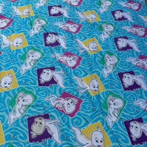 Op de afbeelding: Blauwe stof met een herhaald patroon van het stripfiguur Casper het Vriendelijke Spook. Elke Casper is omlijst in een kleurrijk vierkant, tegen een golvende blauwe achtergrond. De vierkanten zijn geel, paars en groen.