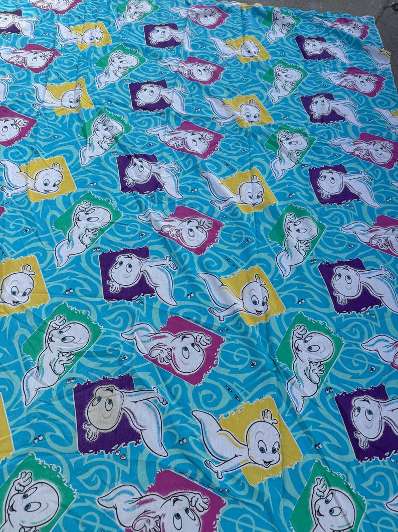 Op de afbeelding: Blauwe stof met een herhaald patroon van het stripfiguur Casper het Vriendelijke Spook. Elke Casper is omlijst in een kleurrijk vierkant, tegen een golvende blauwe achtergrond. De vierkanten zijn geel, paars en groen.