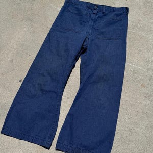 May include: A pair of dark blue denim pants featuring a wide-leg silhouette, a button-fly closure, belt loops at the waistband, and two large, square-shaped patch pockets positioned on the front of the garment.
