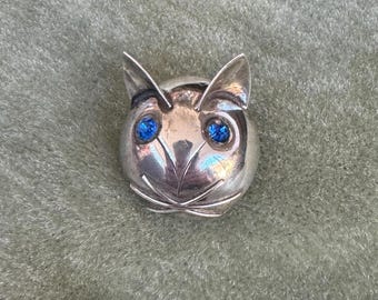 以下が含まれることがあります： 明るい青色の宝石の目を持つ、銀色の猫の形をしたブローチ。 ブローチは尖った耳と様式化された顔をしています。 猫の顔は滑らかで反射性があり、シンプルでエレガントなデザインです。