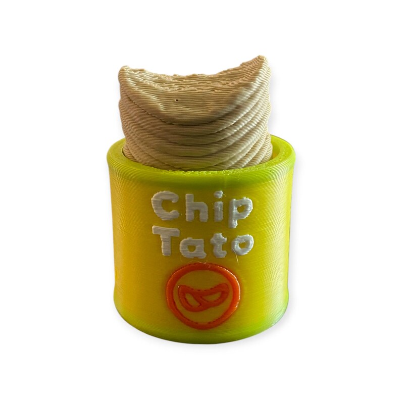 K&ouml;nnte beinhalten: Ein 3D-gedrucktes Novelty-Item, das einem Chip-Beh&auml;lter &auml;hnelt. Der Beh&auml;lter ist gelb mit den wei&szlig;en Worten "Chip Tato". Die Oberseite des Beh&auml;lters ist mit einem strukturierten, beigefarbenen Material gef&uuml;llt, und ein rotes Chip-Logo ist sichtbar.
