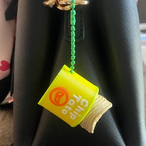 K&ouml;nnte beinhalten: Ein gelb-oranger Miniatur-Chipst&uuml;ten-Anh&auml;nger mit dem Text "Chip Tato" und einem herausragenden Chip. Der Anh&auml;nger ist an einer schwarzen Handtasche mit einem goldenen Verschluss &uuml;ber eine gr&uuml;ne Perlenkette befestigt.