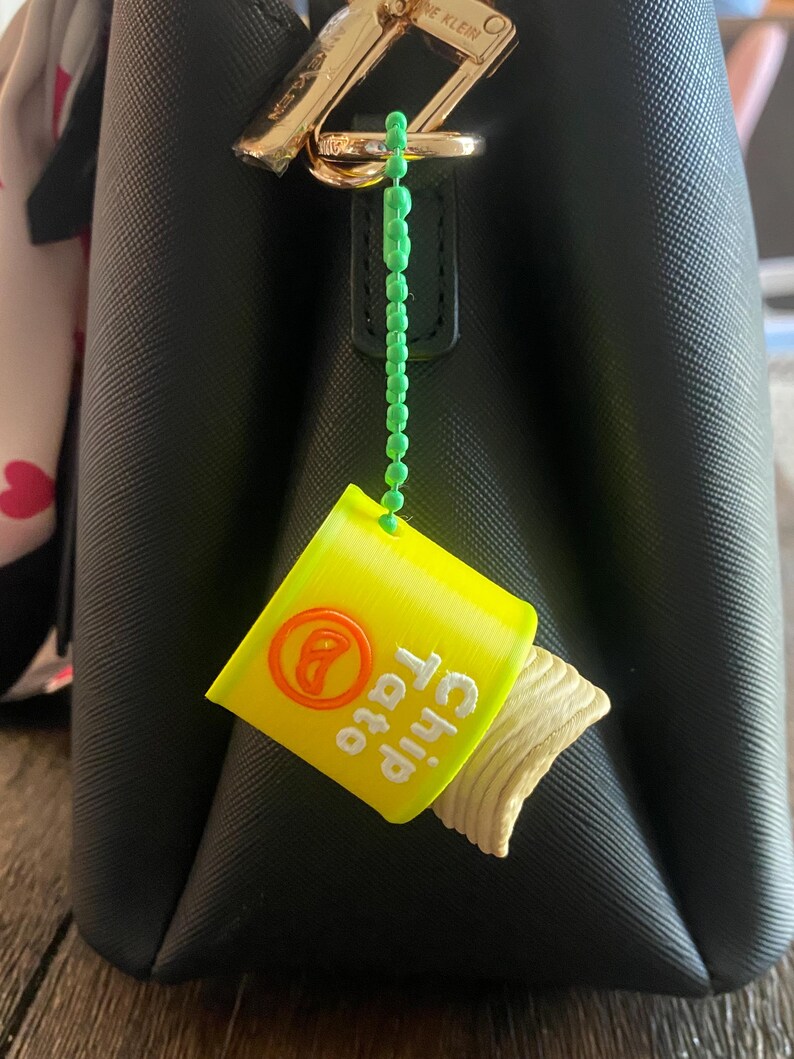 K&ouml;nnte beinhalten: Ein gelb-oranger Miniatur-Chipst&uuml;ten-Anh&auml;nger mit dem Text "Chip Tato" und einem herausragenden Chip. Der Anh&auml;nger ist an einer schwarzen Handtasche mit einem goldenen Verschluss &uuml;ber eine gr&uuml;ne Perlenkette befestigt.