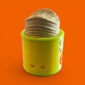 K&ouml;nnte beinhalten: Ein zylindrischer Beh&auml;lter aus dem 3D-Druck in leuchtendem Gelb, der einen Stapel hellbeige, geschichteten Materials enth&auml;lt. Der Beh&auml;lter hat zwei kleine, kreisf&ouml;rmige Aussparungen und ein teilweises Logo in Orange. Der Hintergrund ist einfarbig orange.