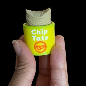 K&ouml;nnte beinhalten: Ein gelber Beh&auml;lter mit dem Text "Chip Tato" in Wei&szlig; und einer rot-orangen Grafik. Der Beh&auml;lter enth&auml;lt einen beigen, zylindrischen Gegenstand. Das Bild ist vor schwarzem Hintergrund.