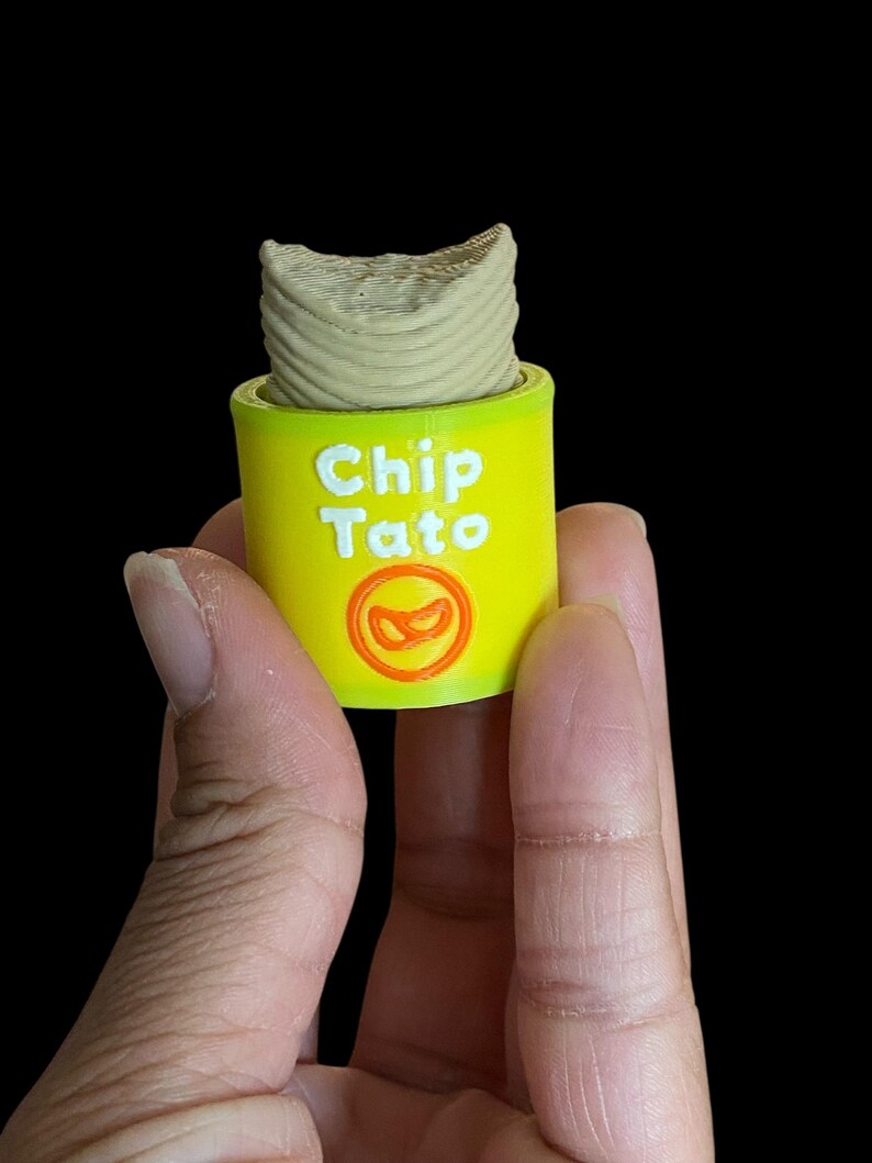 K&ouml;nnte beinhalten: Ein gelber Beh&auml;lter mit dem Text "Chip Tato" in Wei&szlig; und einer rot-orangen Grafik. Der Beh&auml;lter enth&auml;lt einen beigen, zylindrischen Gegenstand. Das Bild ist vor schwarzem Hintergrund.