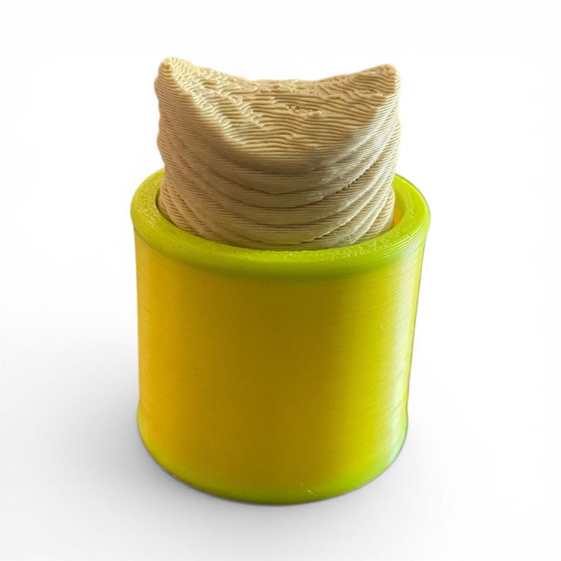 K&ouml;nnte beinhalten: Ein 3D-gedrucktes, zylindrisches Objekt mit einem leuchtend gelben Sockel und einer hellbraunen, strukturierten Oberseite. Der obere Teil hat ein geschichtetes, wellenartiges Muster. Das Objekt steht vor einem wei&szlig;en Hintergrund.